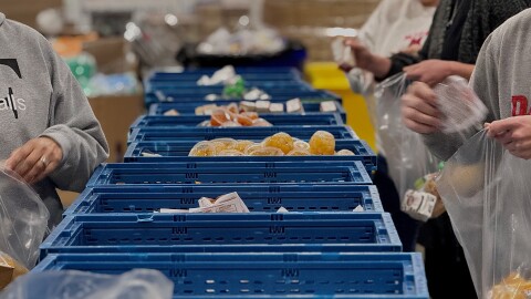 Staff at The Foodbank Inc. stand on either side of blue crates, filled with non-perishable items, to pack boxes 