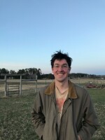 Malachy Koons standing in front of a fence, smiling. He has dark hair and glasses.