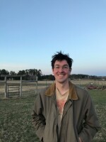 Malachy Koons standing in front of a fence, smiling. He has dark hair and glasses. 