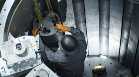 A person wearing gloves and a helmet touches a small cylinder, surrounded by metal-looking walls.