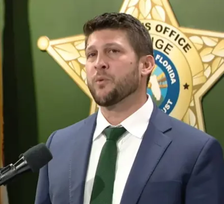 Man with brown hair and a beard, and blue suit, standing behind a microphone with a sheriff's badge behind him