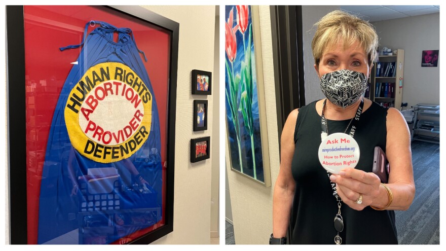 At the left: a framed cape with the words "Human Rights Defender, Abortion Provider" hangs on a wall. At the right: a woman wearing a mask holds a button that reads: "Ask Me How to Protect Abortion Rights"