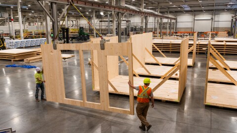 Workers in yellow hardhats and orange vests construct modular timber homes in a warehouse.