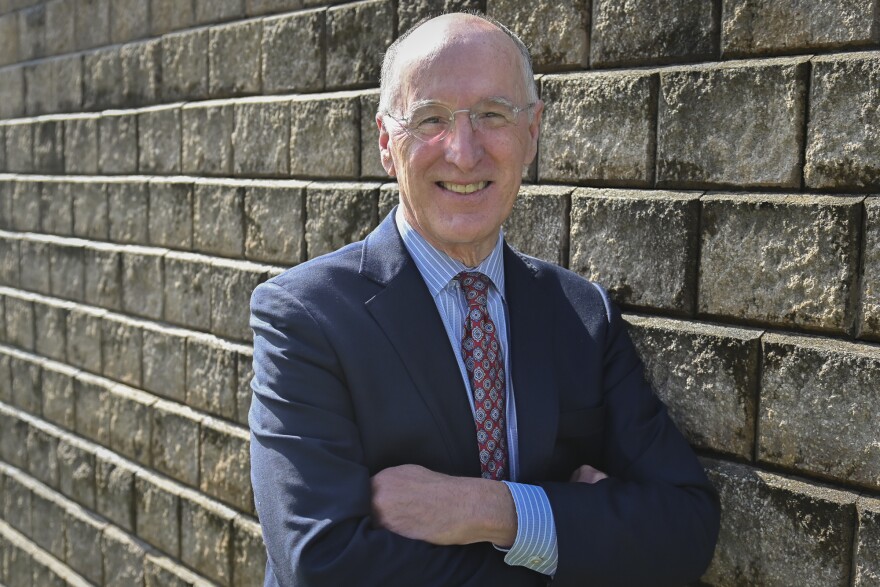 A man wearing a blue suit and red tie leans on a brick wall with his arms folded. He is smiling and looking at the camera.