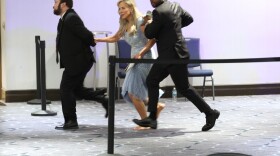 two men in black suits and a woman in a light blue dress running in a hallway. the woman is not wearing shoes