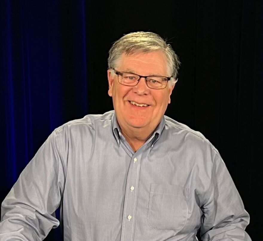 A man with a grey shirt and glasses smiles at the camera.