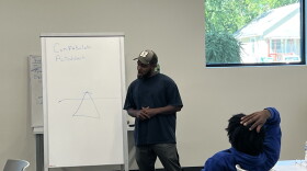 Phillip Townsend stands in front of a classroom. He wears a black t-shirt and dark pants. The classroom has white walls. In this photo, Phillip is speaking to a young man, who is sitting in a chair with a dark blue sweatshirt. 