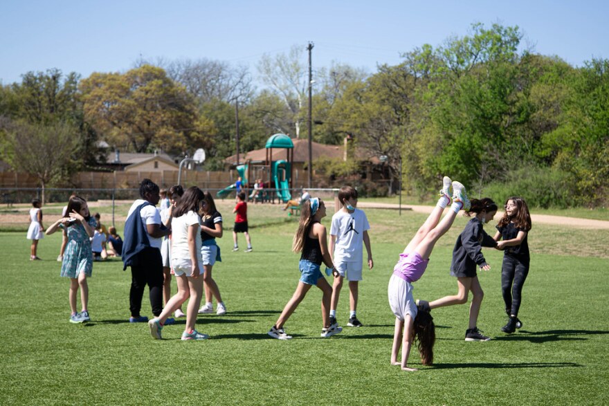 Children play outside on a green field. One girl does a handstand.