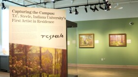 One room of the "Capturing the Campus: T.C. Steele, Indiana University’s First Artist in Residence" gallery space. The a banner is in the center of the room, and Steele's paintings are hung on the wall. 