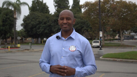 A man with a tennis-ball-sized-pin on his shirt that reads "CALL KEVIN!," stands in a parking lot with his hands together at his torso
