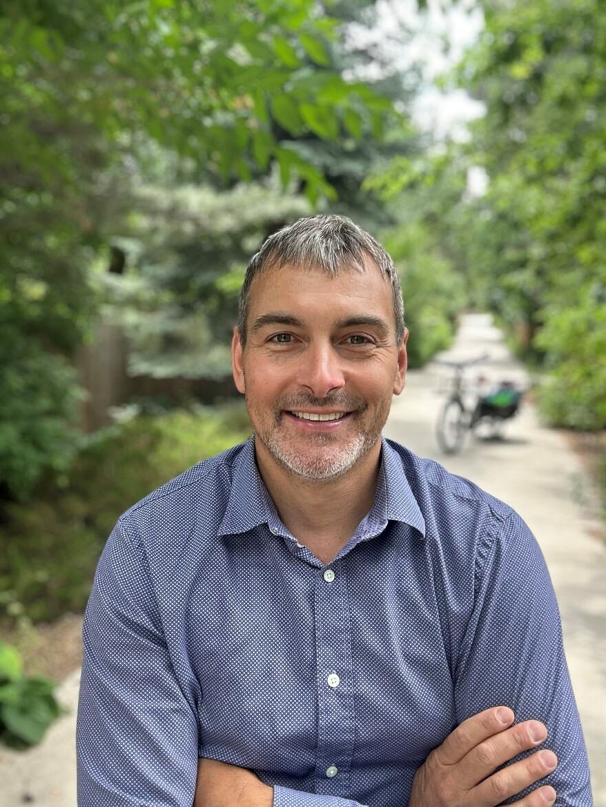 Boulder City Council member Ryan Schuchard, a man in a blue checked shirt and short dark hair stands with his arms folded on a bike path. "The question generally isn't, 'do we have too much parking?' The question is, 'why is it so hard to build more housing, and why don't we have better-functioning transit and more more bikeways?'" says Boulder City Council member Ryan Schuchard, who is part of a group advocating for the city to eliminate its minimum parking requirements for builders and developers.