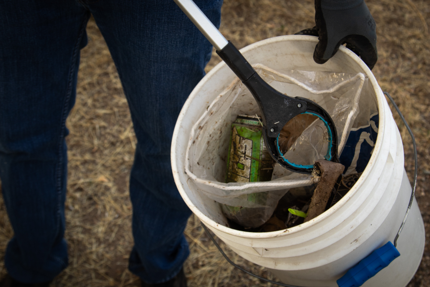 Close-up on a bucket of trash, including an energy drink, all covered in pond muck. A person wearing gloves, mostly off screen, is holding the bucket and reaching into it with a grabber.