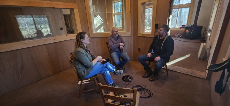 Three people sit in chairs in a wood paneled room. Behind them is a larger room with a wood stove in the corner. On the left is a woman who appears to be interviewing two men on the right. Microphones and cables can be seen.
