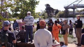 A group of about 30 people standing outside in the sun in front of the water at Berkeley Aquatic Park. Most people's backs are to the camera, but a few are facing the camera, some people are sitting in the dirt. Their heads are cocked and you can tell that they are listening to Wholly H2O tour guide Chris James, who is standing on a bench so people can hear him more clearly. James has a full head of white hair and is wearing a white and blue button down shirt, and blue jeans. He is holding several pieces of paper in his left hand. Many people are wearing hats, and sunglasses. In the background, you can see a bridge, small oak trees, willow, and sage bushes. 