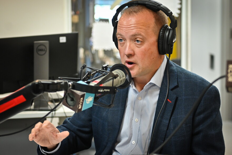 A man wearing a blue sports coat  is wearing a set of headphones, sitting inside a radio studio. He's talking and gesturing with his right hand.