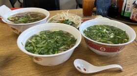 Three bowls of pho sit on a wooden tabletop with a plate of sprouts nearby.