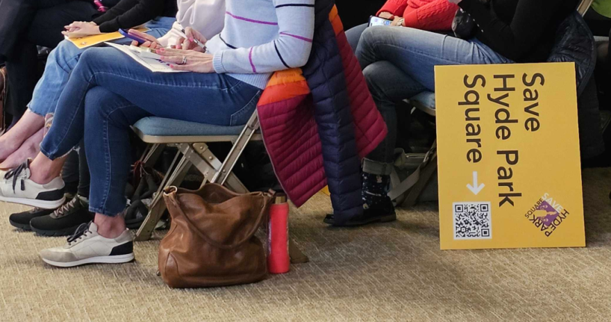 a yellow sign that reads "save hyde park square" stands on its side beside a folding chair where people are seated, photographed from the waist down