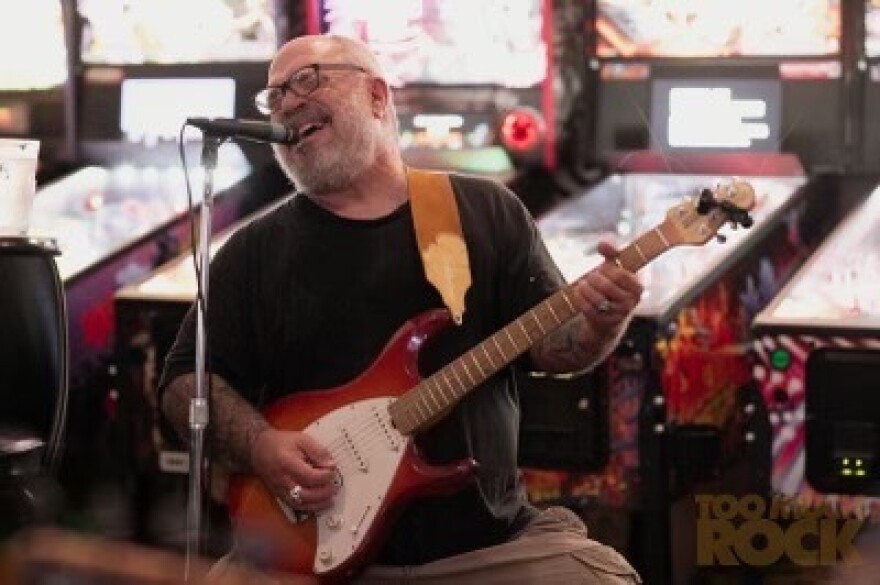 A man in a black tshirt plays guitar and sings into a microphone. Arcade games are behind him