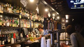 A man leans on a bar while a woman bartender works near rows of bottles of alcohol.