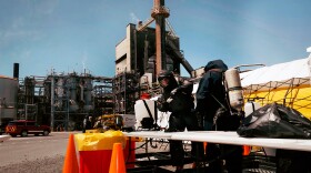 Two people in protective gear with tanks on their back stand with an industrial site in the distance.