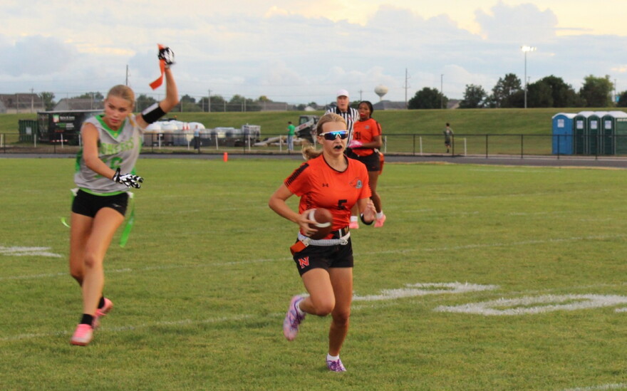 A girls flag football player in an orange and black uniform running with a football while an opposing player chasing them from behind in a gray, green and black uniform holds up a strip of orange tape