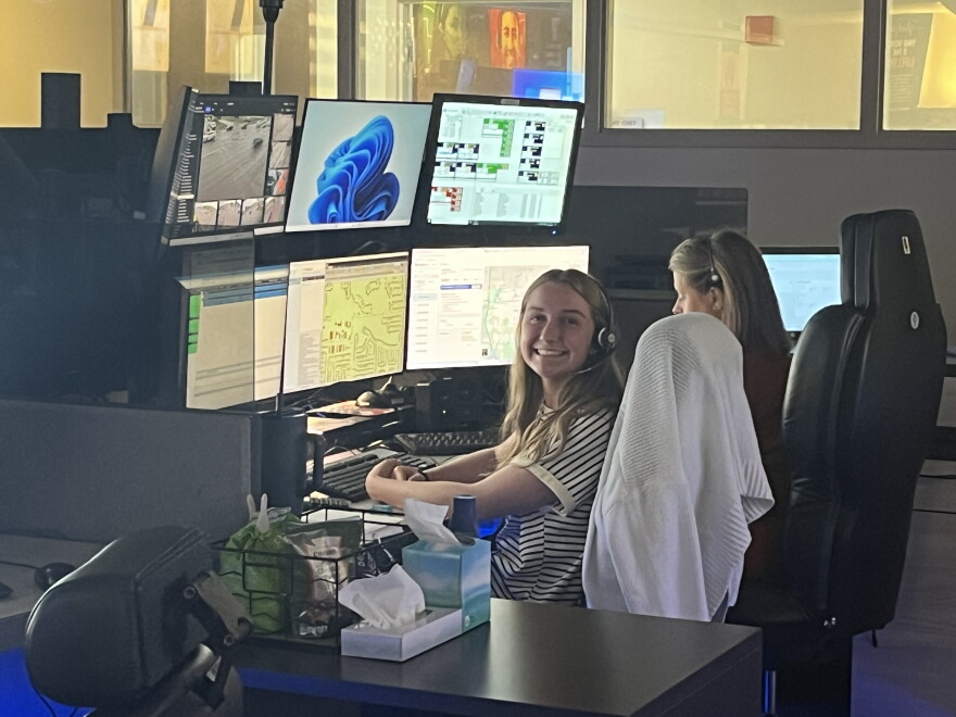 A smiling female emergency dispatcher sits in front of a keyboard and bank of screens at the Bloomington Emergency Dispatch Center.