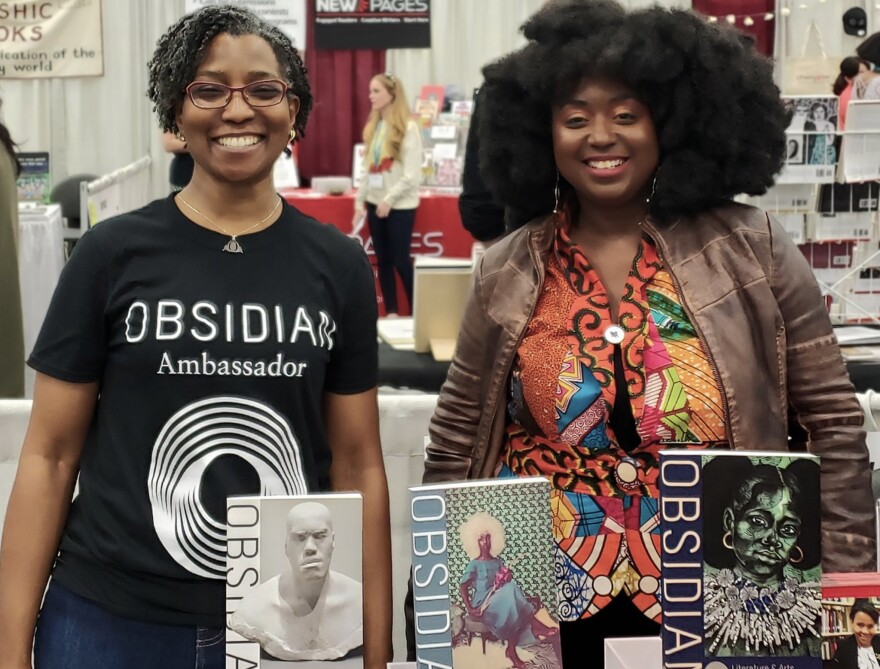 Two women stand smiling behind a table displaying "Obsidian" books with striking cover art at a convention or book fair. One wears an "Obsidian Ambassador" shirt, the other a colorful patterned jacket.