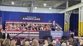 Vice President JD Vance speaks at a podium before a large audience seated in an industrial venue. Red, white, and blue decorations surround the stage, with signs reading “Protecting Americans.” A teleprompter stands beside Vance, and security personnel and supporters are visible in the scene.