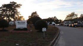 The entrance to Holly Plaza apartments at sundown. There's a bumpy concrete road leading into the apartment complex lined with cars. On the left there's a white picket sign. The sign has black block letters on it reading "Holly Plaza Apartments." The sun is setting behind it. 