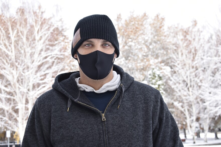 A man in a black mask and beanie from the chest up. The trees behind him are covered by snow.