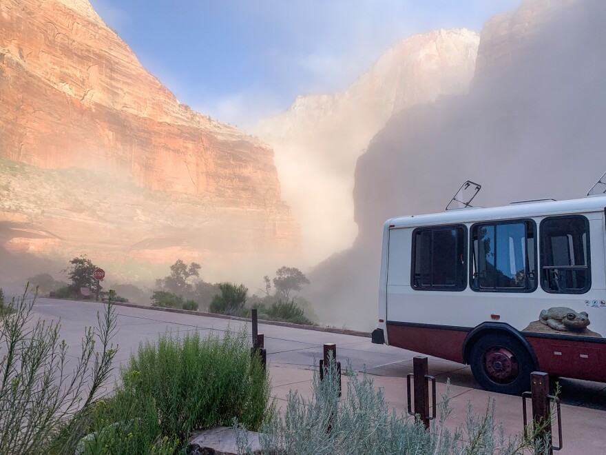 Photo of the rear-end of a Zion National Park Shuttle on a winding canyon road. Clouds of sand that are several times its height billow above. 