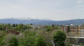 A wide view of Missoula, Montana, shows leafy green neighborhoods and low-rise buildings beneath snow-capped mountains under a hazy spring sky.