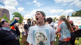 A man in a backwards baseball cap and a Kurt Cobain shirt yells in the center foreground of the photo, amid a crowd of people gathered on the grass near Country Club Plaza. 