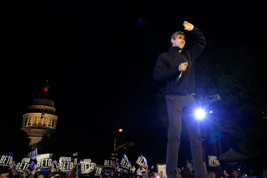 Beto O'Rourke stands on a stage at night, with spotlights lighting him. The state Capitol is lit in the background.