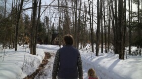 Potato farmer, Amanda Wolters holds her daughter's hand as they walk around their farm and look at damaged trees. Above them is a tree damaged so badly that it's arched over the walkway. The trails are snowy and the sky is blue. 
