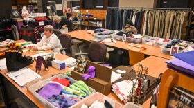 One woman sits at a long table talking. Another woman behind her bends to pick up some material from the floor. They are surrounded by clothes hanging on racks, clothing in baskets and other household items arranged on long tables.
