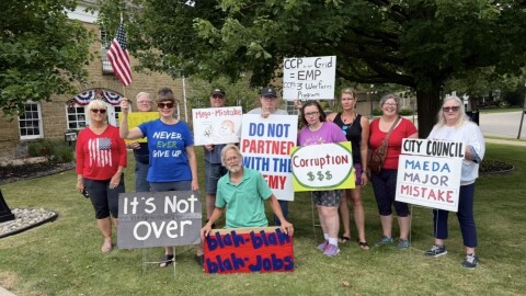 On a green lawn, 10 members of the committee stand with signs in front of a colonial-style building. Their signs talk about corruption and have messages like "City Council MEADA major mistake".