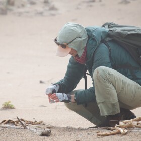 A woman crouches in the sand holding a small cup of water and a tiny egg.