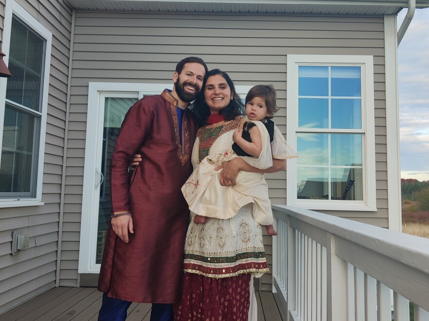 A couple and a baby in traditional Indian dress stand on a porch in front of a cloudy sky.