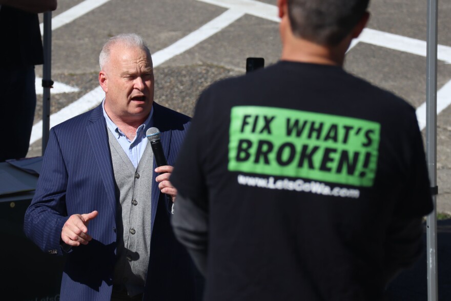 Jim Walsh is seen on the left side of the photo holding a microphone. There's a person in the foreground of the photo wearing a black shirt with a green rectangle, with the phrase "fix what's broken!"