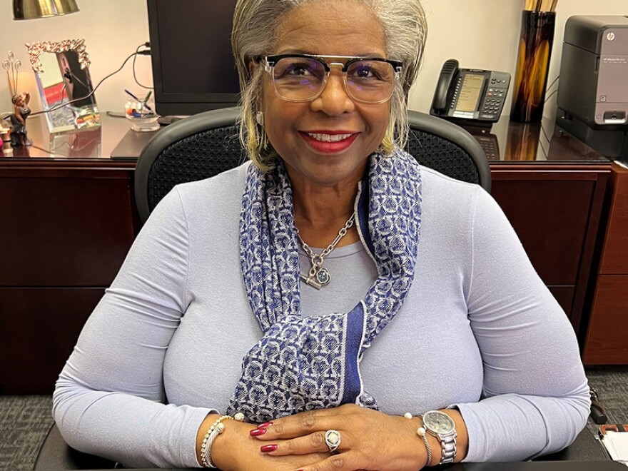 Jeanette Abraham sits at a desk, wearing a blue shirt and blue and white scarf