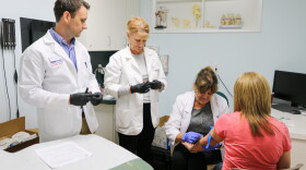 Two standing researchers in lab coats. One researchers is sitting and taking blood from the arm of a blonde patient with their back turned to the camera. 
