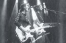 A black-and-white image of a woman playing electric guitar while singing loudly into the microphone on stage at a darkened music venue with spotlights overhead.