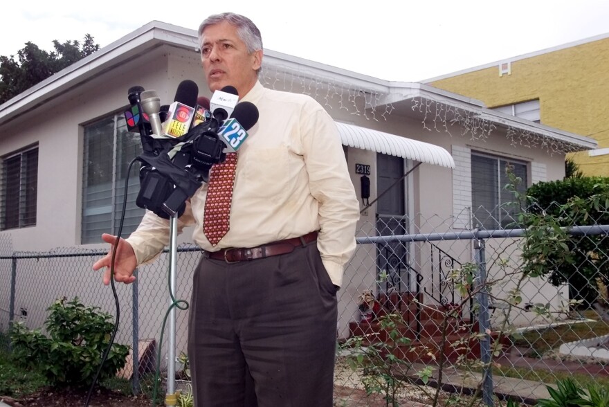 A man speaks into a number of microphones as he stands outside a house