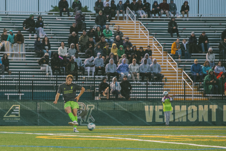 A photo of a soccer player kicking a ball, with people watching in stands in the background. 