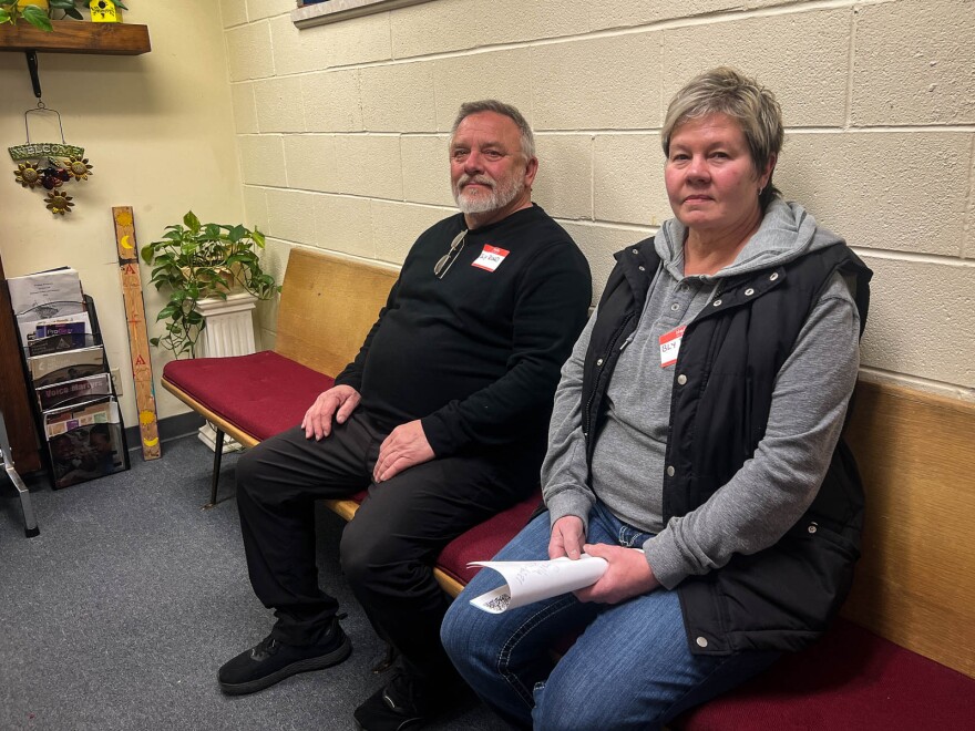 A man and woman sit side by side on a church pew against a wall.