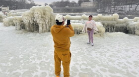 A person poses for a photo in front of frozen bushes. The person taking the picture is in the foreground, wearing all yellow. Everything around them is covered in ice. 