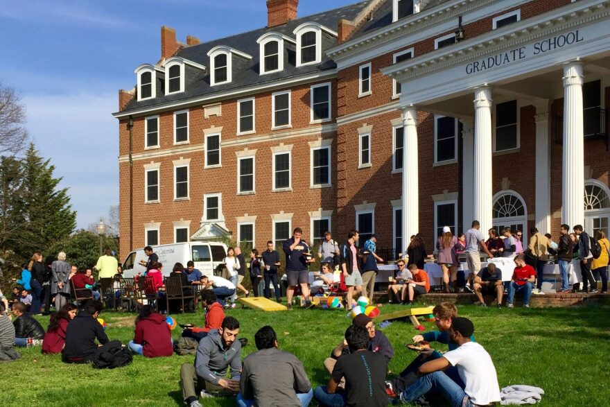 Students sit and stand on a lawn, eating and talking. 