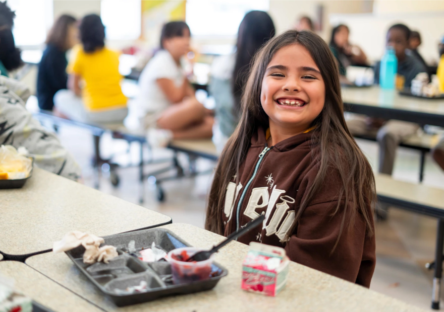 Una niña mira a la cámara con una gran sonrisa, junto a un plato con fruta y otros alimentos.