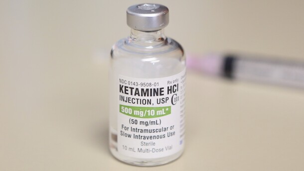 A clear bottle with a label that reads "ketamine" stands on a white table.
